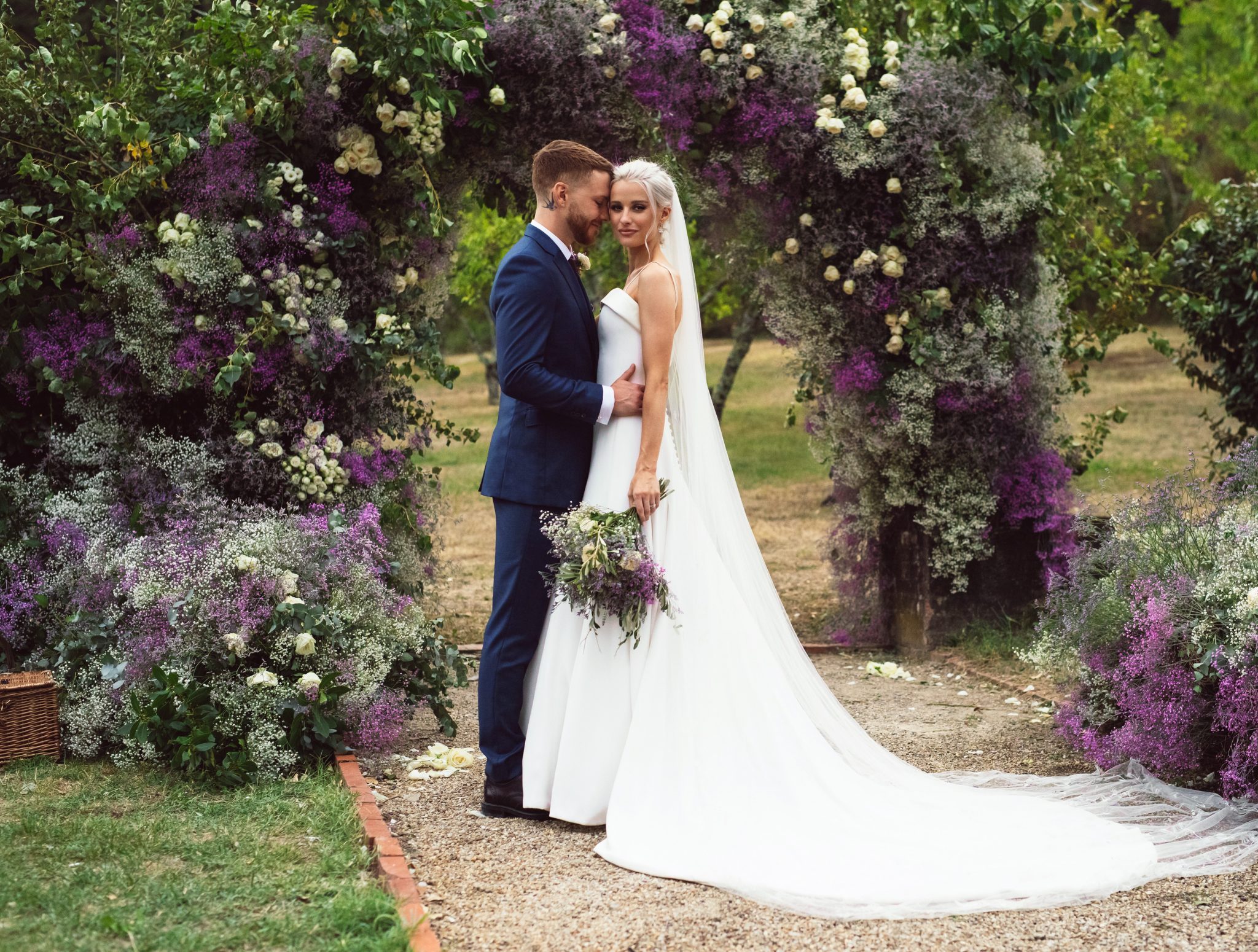 Victoria Magrath of inthefrow stands in front of a flower arch with her husband in a Phillipa Lepley dress carrying her bouquet.